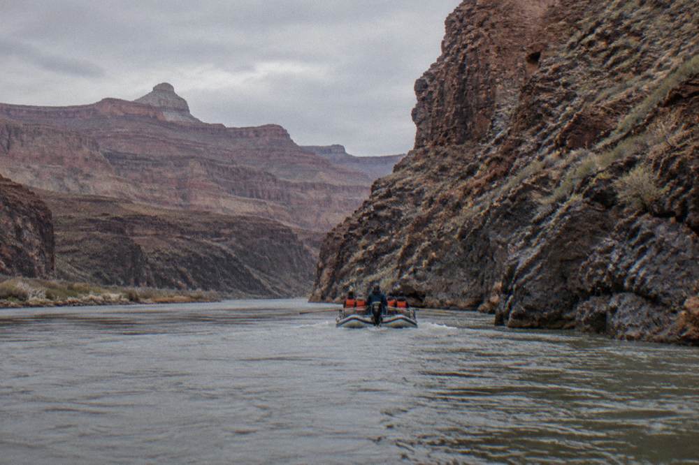 A motorized raft holding paddlers in orange life vests mid-river beneath towering rock cliffs